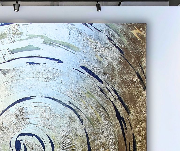 Nahaufnahme einer abstrakten, kreisförmigen Wandkunst mit goldener und blauer Struktur, beleuchtet von zwei schwarzen Deckenspots.